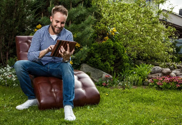 Jeune homme beau en vêtements décontractés assis dans un canapé de luxe avec iPad dans le jardin d'été . — Photo