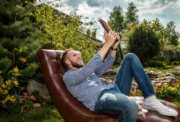 Jeune homme beau en vêtements décontractés assis dans un canapé de luxe avec iPad dans le jardin d'été . — Photo