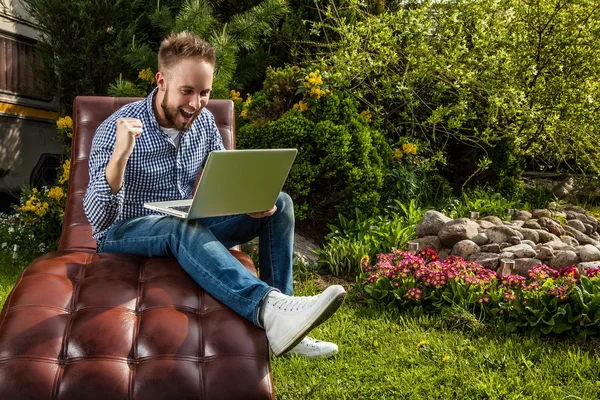 Joven hombre guapo en ropa casual sentarse en un sofá de lujo con portátil en el jardín de verano . — Foto de Stock