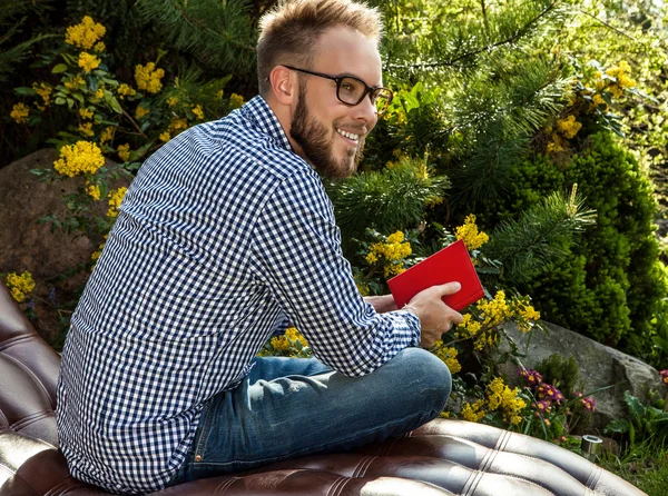 Jovem homem bonito em roupas casuais e óculos sentar no sofá de luxo no jardim de verão . — Fotografia de Stock