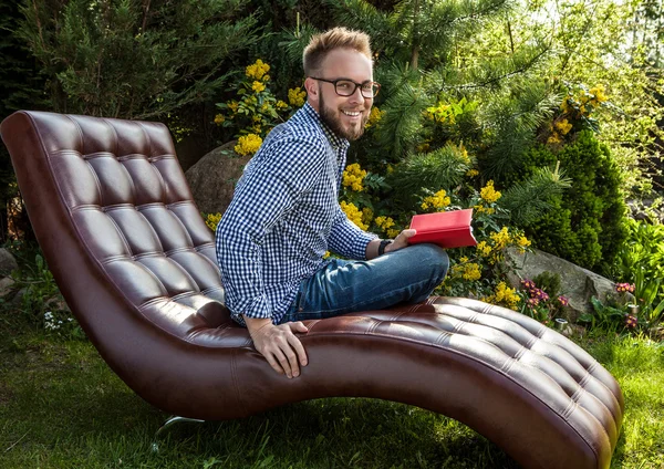 Junger gutaussehender Mann in lässiger Kleidung & Brille sitzt auf Luxus-Sofa im Sommergarten. — Stockfoto