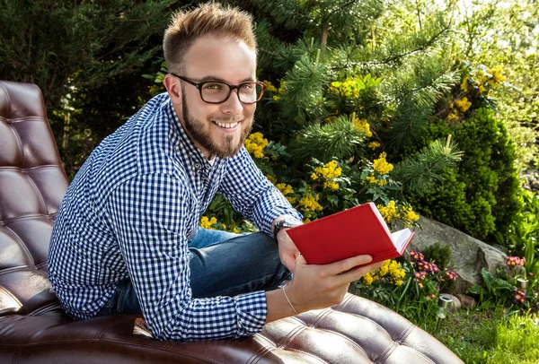 Joven hombre guapo en ropa casual y gafas sentarse en un sofá de lujo en el jardín de verano . — Foto de Stock