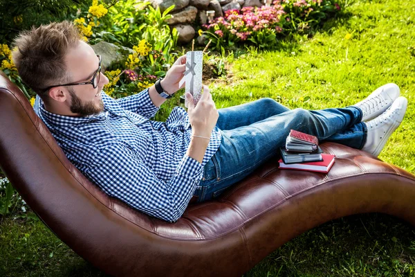 Jeune homme beau en vêtements décontractés assis dans un canapé de luxe avec carte cadeau dans le jardin d'été . — Photo