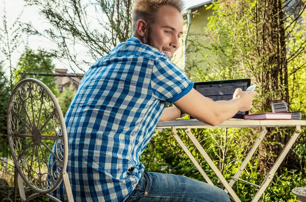 Joven hombre guapo sonriente en ropa casual de trabajo en una mesa de hierro con ordenador contra el jardín del campo . — Foto de Stock