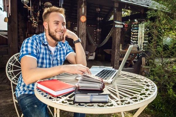 Young smiling handsome man in casual clothes work at an iron table with computer against country garden. — Φωτογραφία Αρχείου