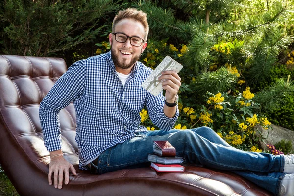 Junger gutaussehender Mann in lässiger Kleidung sitzt auf Luxus-Sofa mit Geschenkkarte im Sommergarten. — Stockfoto