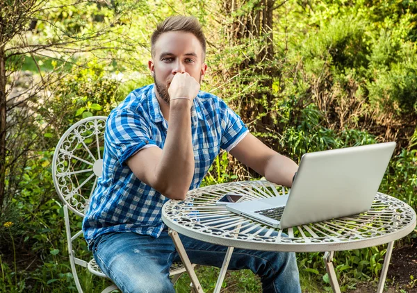 Young smiling handsome man in casual clothes work at an iron table with computer against country garden.