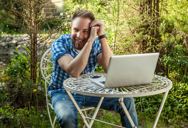 Jovem sorrindo homem bonito em roupas casuais trabalhar em uma mesa de ferro com computador contra jardim de campo . — Fotografia de Stock
