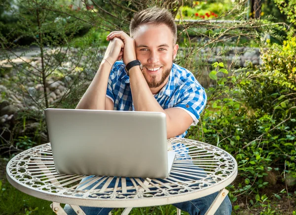 Young smiling handsome man in casual clothes work at an iron table with computer against country garden. — Stock Photo, Image