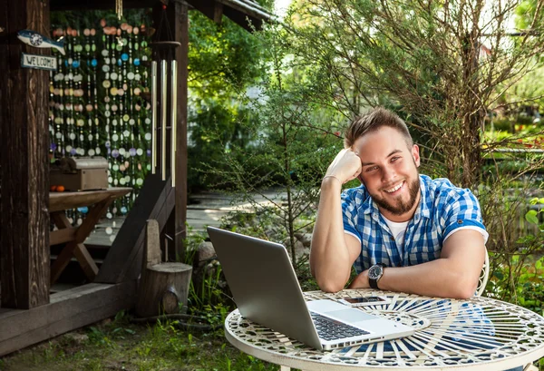 Joven hombre guapo sonriente en ropa casual de trabajo en una mesa de hierro con ordenador contra el jardín del campo . — Foto de Stock