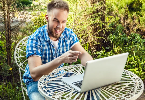 Giovane uomo bello sorridente in abiti casual lavorare a un tavolo di ferro con il computer contro giardino di campagna . — Foto Stock