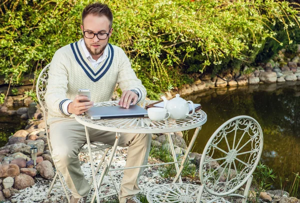 Jovem elegante homem bonito em roupas confidenciais brilhantes trabalhar na mesa de ferro com computador e bule de chá contra o jardim do país . — Fotografia de Stock