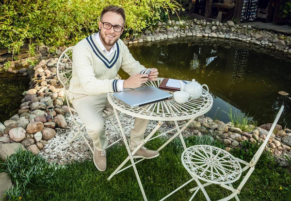 Young stylish handsome man in bright confidential clothes work at iron table with computer & teapot against country garden. — Stok fotoğraf