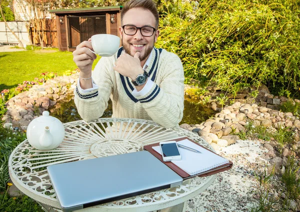 Young stylish handsome man in bright confidential clothes work at iron table with computer & teapot against country garden. — Stockfoto