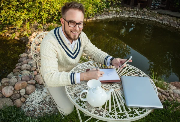 Young stylish handsome man in bright confidential clothes work at iron table with computer & teapot against country garden. — Zdjęcie stockowe