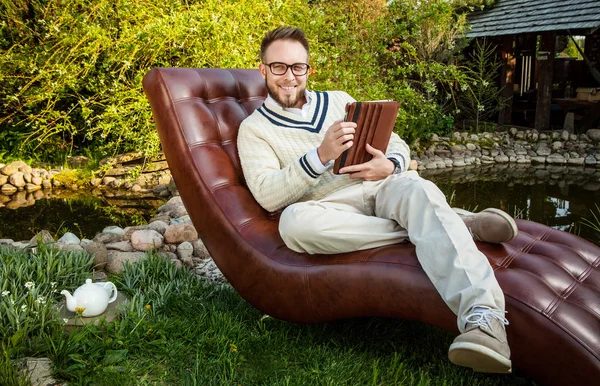 Young handsome man in glasses sit in luxury sofa with iPad in summer garden. — Stockfoto