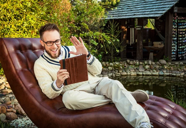 Junger gutaussehender Mann mit Brille sitzt auf Luxus-Sofa mit Ipad im Sommergarten. — Stockfoto