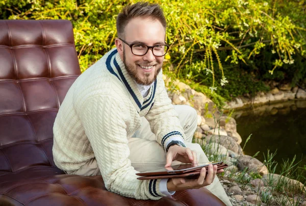 Young handsome man in glasses sit in luxury sofa with iPad in summer garden. — Stok fotoğraf