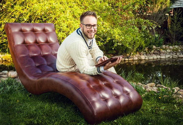 Jeune bel homme en lunettes assis dans un canapé de luxe avec iPad dans le jardin d'été . — Photo