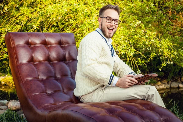 Young handsome man in glasses sit in luxury sofa with iPad in summer garden. — Stock fotografie