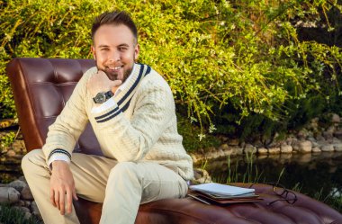 Young handsome man in glasses sit in luxury sofa with iPad in summer garden.