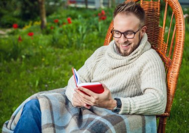 Handsome man relax in rocking-chair & reading red book in summer garden.