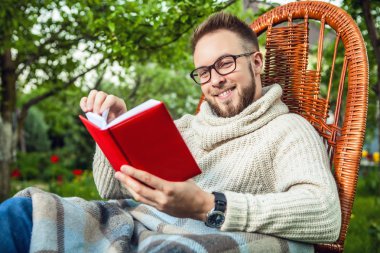 Handsome man relax in rocking-chair & reading red book in summer garden.