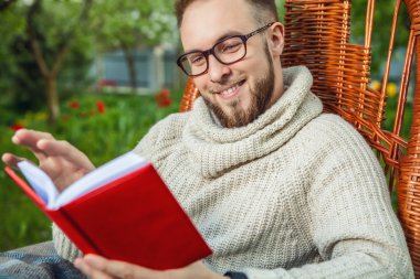 Handsome man relax in rocking-chair & reading red book in summer garden.