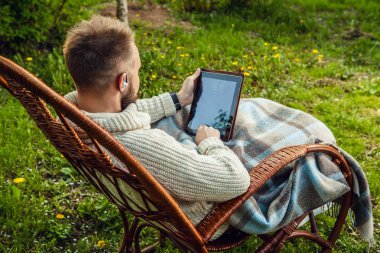 Friendly man sits in a rocking-chair with plaid & tablet in summer country garden.