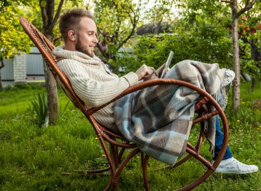Friendly man sits in a rocking-chair with plaid & tablet in summer country garden.