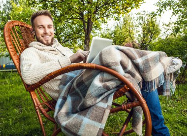 Friendly man sits in a rocking-chair with plaid & tablet in summer country garden.