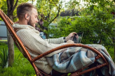 Friendly man sits in a rocking-chair with plaid & tablet in summer country garden.