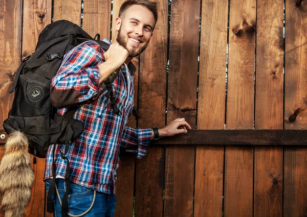Joven hombre guapo turista viajero con una mochila de marcha posa contra valla de madera . —  Fotos de Stock