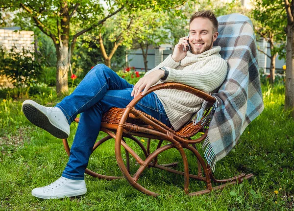 Schöner Mann relaxt im Schaukelstuhl mit Plaid & Telefon im Sommergarten. — Stockfoto