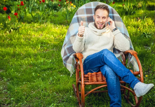 Bello uomo rilassarsi in sedia a dondolo con plaid & telefono in un giardino estivo . — Foto Stock