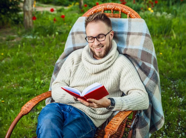 Schöner Mann entspannt sich im Schaukelstuhl & liest rotes Buch im Sommergarten. — Stockfoto