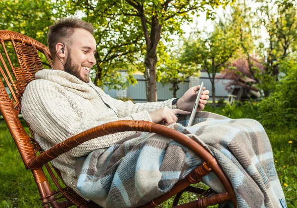 Friendly man sits in a rocking-chair with plaid & tablet in summer country garden. — Stockfoto