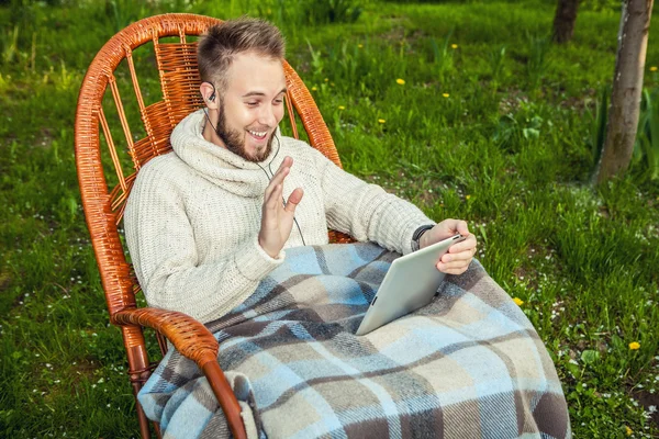 Amistoso hombre se sienta en una mecedora con cuadros y tabletas en el jardín del país de verano . — Foto de Stock