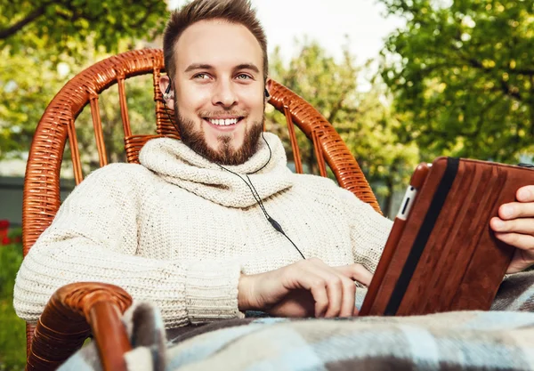 Amistoso hombre se sienta en una mecedora con cuadros y tabletas en el jardín del país de verano . — Foto de Stock