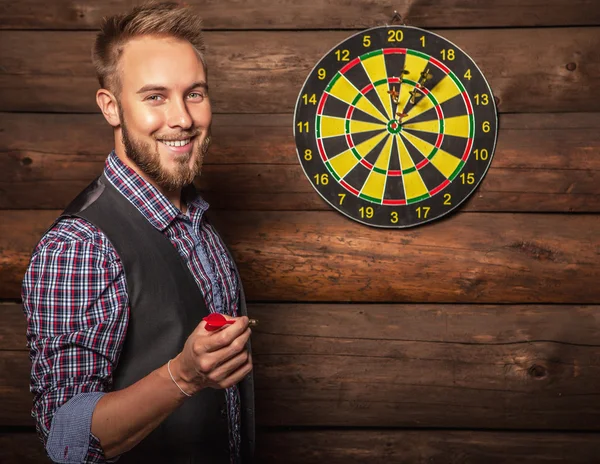 Portrait of young friendly lucky man against old wooden wall with darts game. Concept: Hit in purpose. Photo. — Stockfoto