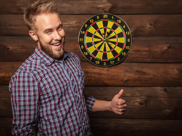 Portrait of young friendly lucky man against old wooden wall with darts game. Concept: Hit in purpose. Photo. — Stock Fotó
