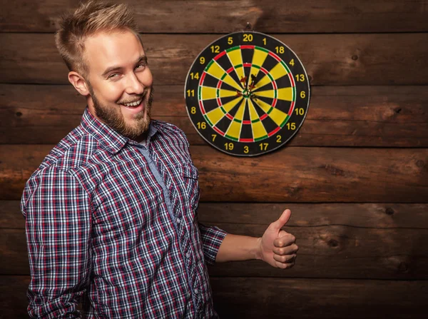 Portrait of young friendly lucky man against old wooden wall with darts game. Concept: Hit in purpose. Photo. — Zdjęcie stockowe