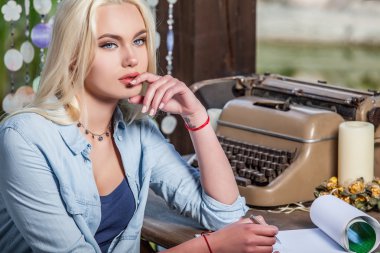 Beautiful young blonde sits in arbor at oak table near vintage typewriter.
