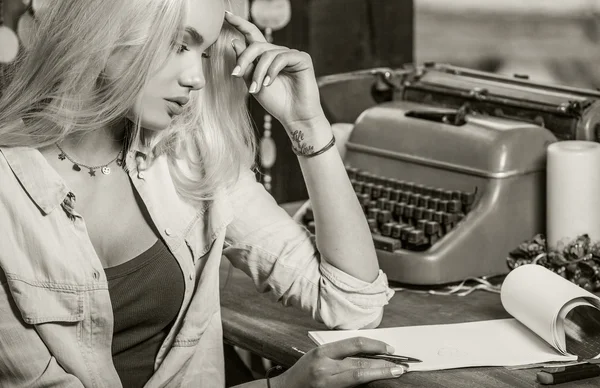 Beautiful young blonde sits in arbor at oak table near vintage typewriter & makes entries in sheets of paper. Sepia photo. — 图库照片
