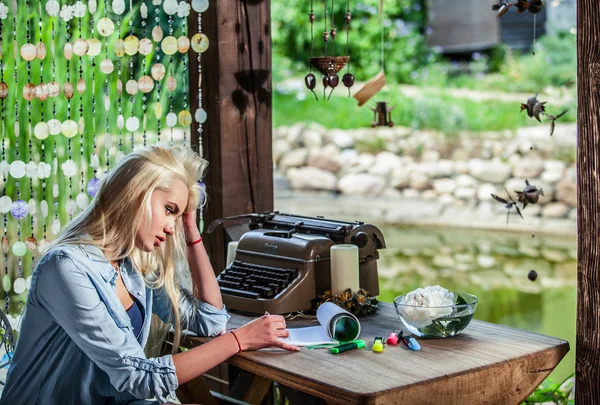 Beautiful young blonde sits in arbor at oak table near vintage typewriter. — Stockfoto