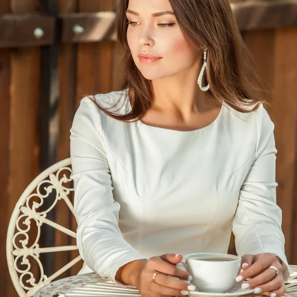 Outdoors portrait of beautiful young girl in luxury white dress posing near garden metal table with cup of coffee. — Stock Fotó