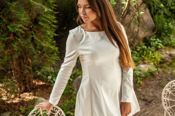 Portrait extérieur de belle jeune fille en robe blanche de luxe posant près de la table en métal de jardin avec tasse de café . — Photo