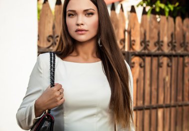 Outdoors portrait of beautiful young girl in luxury white dress posing near gate to country house in sunny summer day.