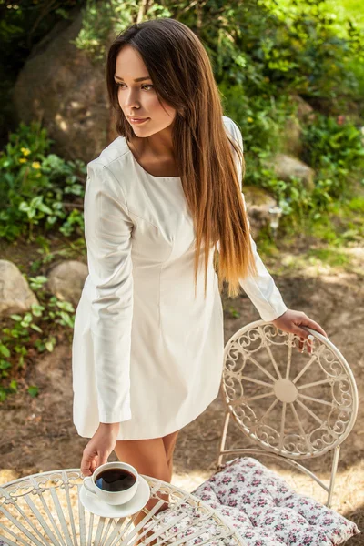 Retrato al aire libre de una hermosa joven en vestido blanco de lujo posando cerca de la mesa de metal jardín con taza de café . —  Fotos de Stock