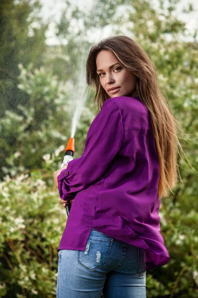 Hermosa joven alegre en camisa casual violeta posa en un jardín de verano con manguera de agua . — Foto de Stock